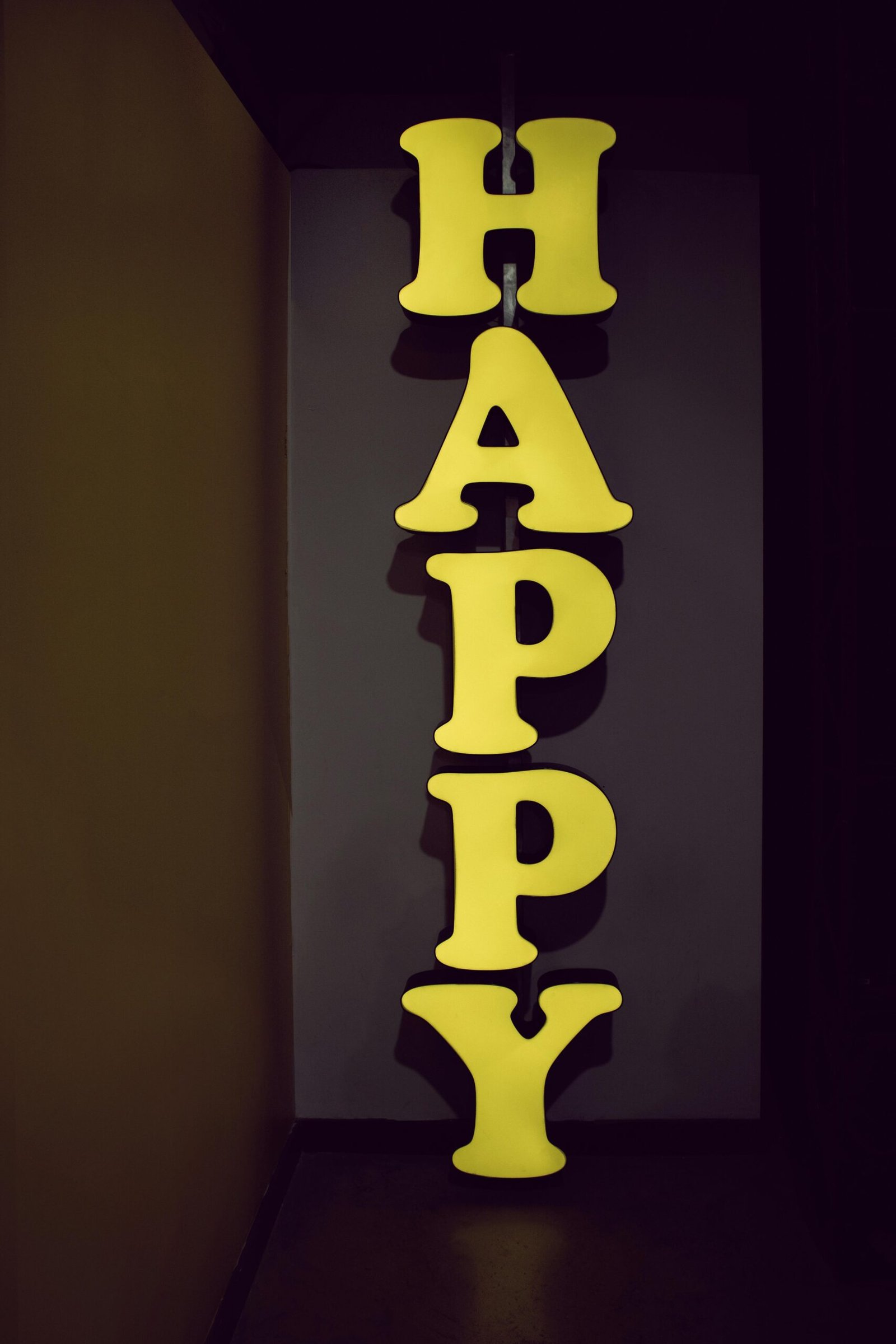 Illuminated yellow 'HAPPY' sign displayed vertically in a dimly lit hallway setting, evoking a playful mood.